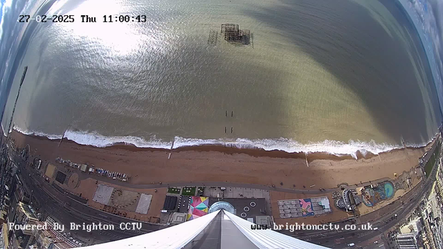 Aerial view of a beach with a smooth sandy shore and gentle waves lapping at it. The water reflects sunlight, creating a shimmering effect. In the distance, a derelict pier is visible above the water, while a small number of people can be seen walking along the beach. The foreground includes patches of vibrant colors from beachside attractions and structures, with a circular feature and some parked vehicles. The top part of the image shows a tall structure, possibly a tower, pointing downward toward the beach. The time and date are displayed in the upper left corner.