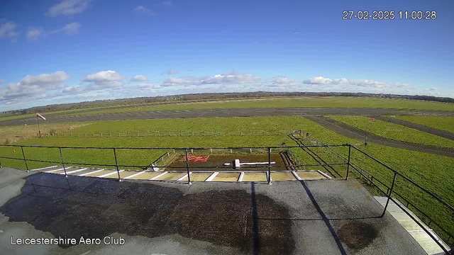 A clear sky with scattered clouds is visible above a green landscape. There is a runway marked with dark asphalt, leading towards the horizon. To the left, there is a red and white windsock indicating the wind direction. In the foreground, a railing encloses a balcony area, while puddles of water hint at recent rain. Fencing lines the grassy areas, indicating an aerodrome environment. The scene captures a serene outdoor setting typical of an airport.