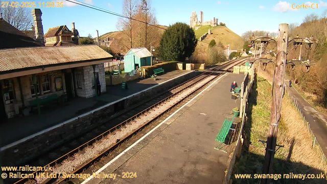 A view of a train station scene from an elevated angle, featuring a platform with two tracks running parallel to each other. The station has a stone building with a sloped roof on the left, and green benches placed along the platform. In the background, a green shed can be seen, along with a small wooden fence. Beyond the station, there are rolling hills topped with ruins of a castle. The sky is clear with a few clouds, and the scene is well-lit, suggesting a sunny day. A couple of people are visible, one tending to a bench and another walking along the path beside the station.