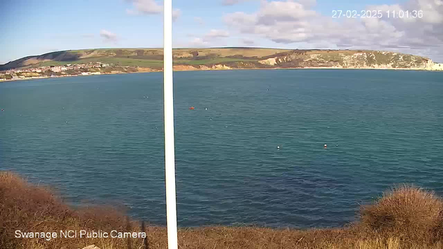 A scenic coastal view featuring a calm blue sea in the foreground and rolling green hills in the background. The shoreline is dotted with a small town, partially visible along the edge, with a mix of buildings. A few small boats can be seen floating on the water, and there are a few white buoys visible as well. The sky is partly cloudy with patches of blue peeking through. In the foreground, there are some brownish bushes or grass. A white pole stands on the left side of the image.