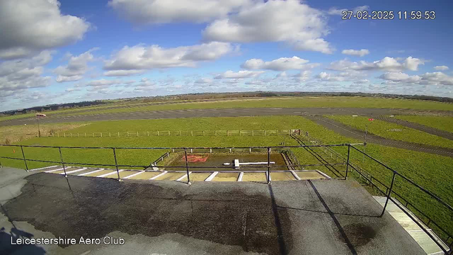 A clear daytime view from a webcam at Leicestershire Aero Club, showing a wide grassy area and a runway. The sky is blue with fluffy white clouds. There is a wooden fence in the middle ground, and a red and white windsock on the right side of the image. A small area of pavement with what appears to be a model airplane is visible in the foreground. The overall scene conveys an open and expansive landscape.