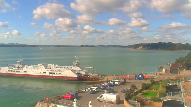 A ferry boat is docked at a pier, with several vehicles parked nearby, including cars and a van. The scene features a calm body of water reflecting the blue sky, which is dotted with fluffy white clouds. In the background, a wooded area and a small coastal cliff are visible, while the waterfront has a mix of greenery and paved surfaces.