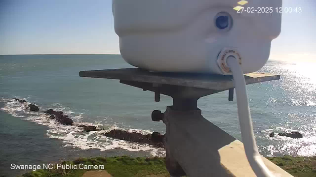 The image shows a view of the ocean from a webcam. In the foreground, there is a white container with a hose attached, resting on a metal platform. Below the platform, rocky formations can be seen emerging from the water. The ocean is calm with gentle waves lapping at the rocks. The sky is clear, and the sun is shining, creating reflections on the water’s surface. The scene is bright and sunny, with greenery visible on the shore.