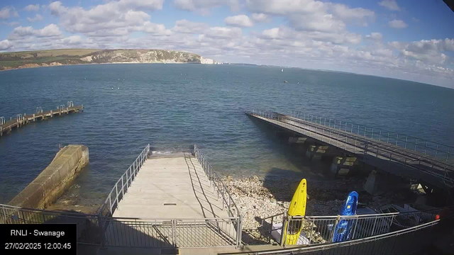 A view from a webcam showing a coastal area with the sea in the foreground. There are two kayaks, one yellow and one blue, positioned on a stony shore next to a wooden platform. The platform extends into the water and is accompanied by several railings. In the distance, white cliffs rise above the shoreline, partially obscured by clouds in a predominantly blue sky. Gentle waves are visible on the surface of the water.