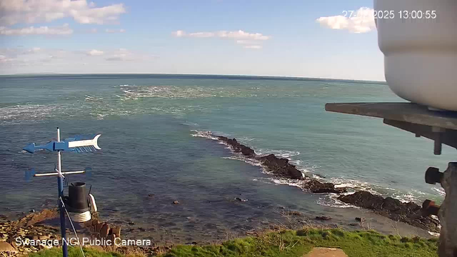 A coastal view captured from a webcam. In the foreground, there is a rocky shoreline leading into the water. A white container is partially visible on the right side of the frame, while a weather vane with a light blue color is attached to a pole. The ocean stretches out into the background, showing gentle waves and a blue sky with scattered white clouds. The sunlight creates a shimmering effect on the water's surface. The image captures an overall serene and natural seaside environment.