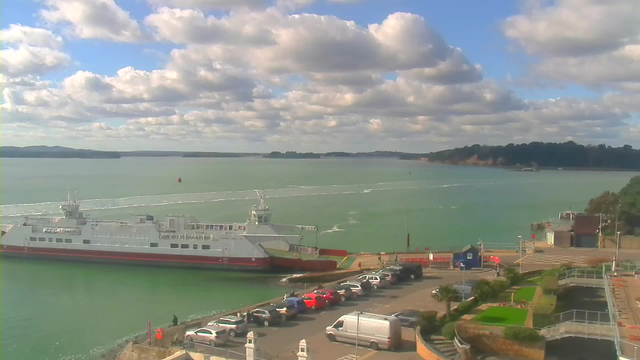 A view of a coastal scene featuring a ferry docked at a port, surrounded by water. The ferry is white with red accents. In the foreground, there is a parking area with several cars, including red and silver vehicles. Lush greenery and a few trees are visible, alongside a series of walkways. The sky is partly cloudy with blue areas peeking through, and hills can be seen in the distance across the water.