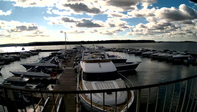 A marina scene featuring multiple boats moored at docks. In the foreground, a large white motorboat is prominently positioned. Several smaller boats surround it, while more boats are seen in the background, floating in calm waters. The sky is partly cloudy with patches of blue and scattered clouds, reflecting sunlight. The water has gentle ripples, indicating a mild breeze. The setting is tranquil, suggesting a peaceful day at the marina.