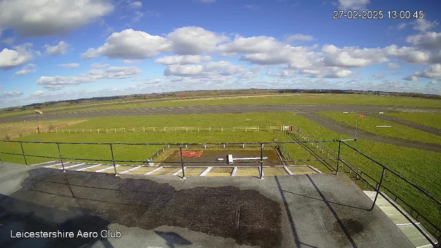 A view from a high vantage point at Leicestershire Aero Club. The foreground features a flat rooftop with railing, and a small wet area visible. In the background, expansive green fields stretch across the scene under a partly cloudy blue sky. There is a tarmac runway to the right, intersecting with the fields, and a small windsock near the edge of the runway indicates wind direction. Beyond the runway, more fields and a distant horizon are visible. The date and time are displayed in the upper right corner.