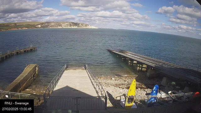 A coastal scene featuring a calm sea under a partly cloudy sky. In the foreground, a wooden pier extends into the water, and there is a small beach area with pebbles. To the right, several brightly colored kayaks are stored near the water's edge. In the background, a shoreline with greenery and cliffs is visible.