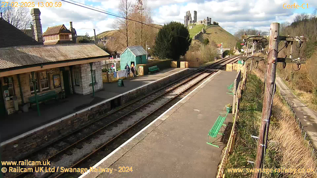 A railway station scene during the day. The platform is made of stone and concrete, with several green benches lining the sides. A small, light blue shed is on the left, and a sign reading "WAY OUT" is placed on the ground. In the background, there are green hills topped with historical ruins. The sky is partly cloudy with some blue peeking through. A few people are walking toward the station, creating a lively atmosphere.