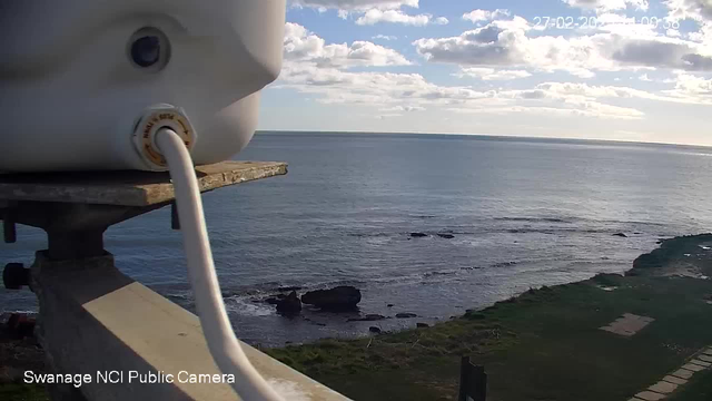 A close-up view of a white cylindrical object connected to a white tube, positioned on a weathered railing. In the background, there is a serene ocean landscape under a partly cloudy sky. The water appears calm with a few rocks visible along the shoreline. The foreground includes patches of green grass.