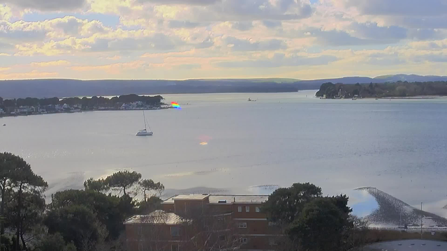 A serene bay scene features a calm body of water reflecting a cloudy sky with shades of blue and gray. In the foreground, a building with a flat roof is partially visible, surrounded by leafy trees. A sailboat is anchored in the water, and a rainbow-colored light can be seen reflecting on the surface. In the distance, a shoreline is lined with houses and distant hills, creating a peaceful coastal atmosphere.