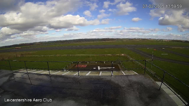 A wide view of an airfield under a cloudy sky. The foreground shows a rooftop surface with a railing, while the middle ground features a grassy area and a runway marked by black asphalt. A few scattered clouds are visible in the blue sky, and there are distant fields and trees on the horizon, indicating a rural setting. The time and location are noted on the image.