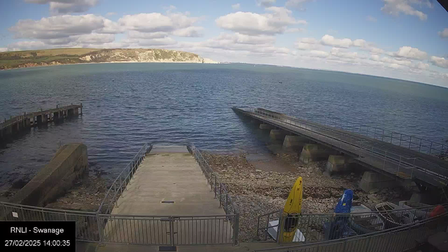 A scenic view of a harbor at low tide shows a concrete pier extending into the calm blue sea. The shoreline is rocky and visible, with patches of light sand. Small boats, including one yellow and one blue kayak, are stored on the beach area next to the pier. In the background, gentle hills with green vegetation rise, leading up to high cliffs that are partially obscured by clouds in the bright sky. The image conveys a serene coastal setting.