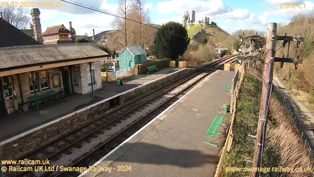 A sunny railway station scene with a stone platform and two sets of railway tracks. On the left, there is a building with a sloped roof, windows, and a green bench outside. In front of the building, there are posters visible on the walls. A green shed is positioned beside the tracks, with benches nearby. The area is lined with wooden fencing, and a hill in the background features a castle ruin. The sky is partly cloudy. The foreground shows a clear view of the tracks leading off into the distance.