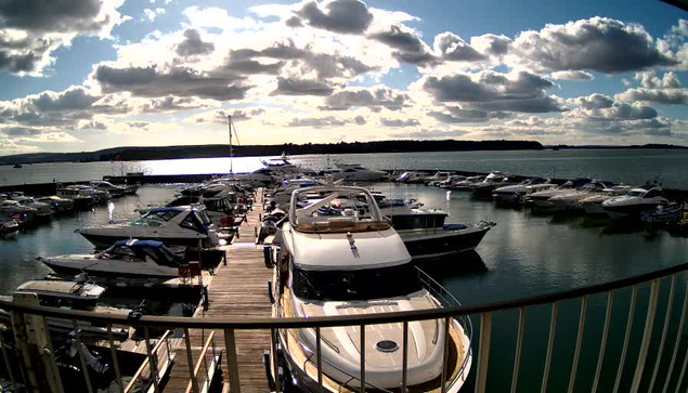 A marina scene with numerous boats docked in calm water. In the foreground, a large white motorboat is prominently positioned. Several smaller boats are scattered around, creating a busy marina atmosphere. The background features a shimmering water surface reflecting the sunlight, along with hills in the distance. Fluffy clouds fill the sky, casting shadows on the water and boats. The scene conveys a tranquil yet lively coastal environment.