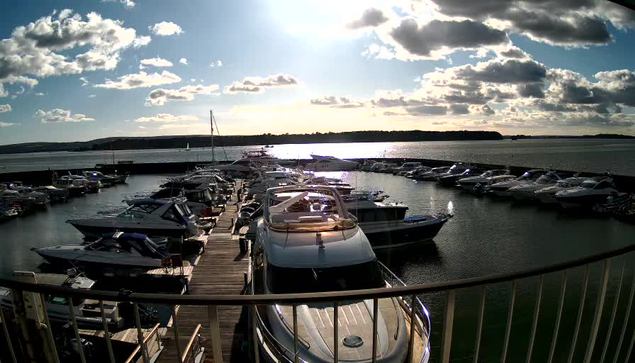 A marina scene under a bright blue sky with scattered fluffy clouds. The sun is shining, reflecting on the water. Numerous boats are docked in the marina, some are large yachts, while others are smaller vessels. A wooden walkway runs along the marina, leading to the boats. The water appears calm, with gentle ripples. In the distance, hills are visible along the shoreline.