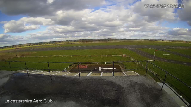 A view from a webcam overlooking an aerodrome on a cloudy day. The foreground features a rooftop railing with a gray surface, leading to a green grassy area. In the distance, there are multiple shades of green fields, a black asphalt runway, and visible wooden fencing along the perimeter. The sky is partly cloudy with varying shades of gray and blue. The timestamp shows the date and time as 27th February 2025, 15:00:08.