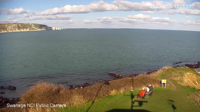 A scenic coastal view featuring the ocean and rocky shoreline. In the foreground, there is a grassy area where three people are sitting on a bench, enjoying the view. The ocean is calm, with a few small waves, and the sky is mostly clear with some fluffy white clouds. In the background, cliffs are visible along the coastline, and there is a signpost near the edge of the grass. The date and time are noted at the top of the image.