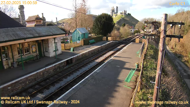 A railway station platform is viewed from an elevated angle. On the left, there is a stone building with a sloped roof and a wooden beam structure, flanked by a green bench. To the right, there are railway tracks leading into the distance, bordered by gravel and grass. A small green shed is visible further down the platform, along with several wooden benches. In the background, a hill rises steeply, topped with the remains of a castle. The sky is mostly clear with a few clouds, indicating daylight.