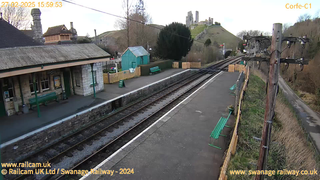 A railway station scene at Corfe with a view of the platform. On the left, there is a stone building with a sloped roof, featuring large windows and a wooden bench in front. A green wooden shed is visible further back, surrounded by trees and a fence. The platform is lined with several green benches and has tracks running alongside it. In the background, a hill rises with the ruins of a castle perched on top, under a clear sky. The image is taken in daylight, showcasing the peaceful setting of the station.