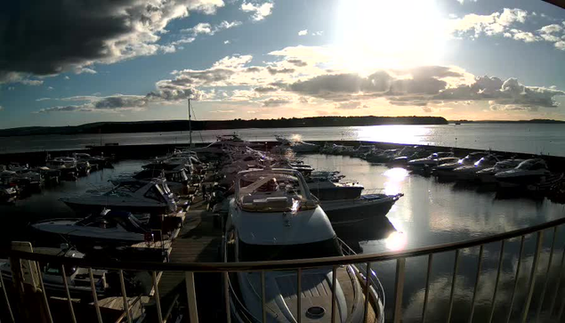 A bustling marina at sunset, with numerous boats moored in calm waters. The sun is low in the sky, casting reflections on the water's surface. Fluffy clouds are visible, and a hill can be seen in the distance. The scene is peaceful, with some boats displaying their silhouettes against the golden light.