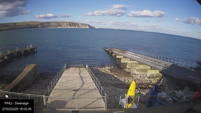 A view of a coastal area featuring a calm blue sea under a partly cloudy sky. In the foreground, there is a wooden pier extending into the water, with several small boats anchored nearby. On the right, a ramp leads down to the water, flanked by rocky shoreline. A bright yellow kayak is visible near the base of the ramp, alongside a blue kayak. The background includes cliffs and green hills extending along the coast, illustrating a scenic landscape.