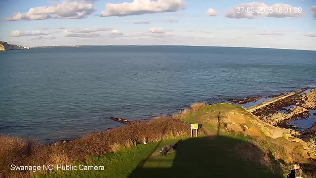 A scenic view of a coastal landscape featuring a calm blue sea under a partly cloudy sky. In the foreground, there is green grass with a pathway leading down. To the right, rock formations create a textured shoreline. A signpost is visible near the edge of the grass, and silhouettes of two people can be seen walking on the pathway. The time stamp in the corner indicates the date and time of the image.