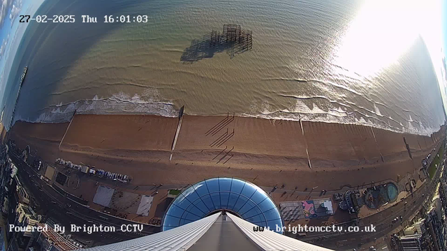 A high view overlooking a sandy beach and the sea below. In the foreground is a circular glass structure, resembling the top of a tower. The beach stretches along the bottom, with people walking and shadows cast on the sand. Waves from the sea gently lap at the shore. To the left, remnants of a pier can be seen emerging from the water. The time and date are displayed in the upper corner, indicating it's a Thursday afternoon.