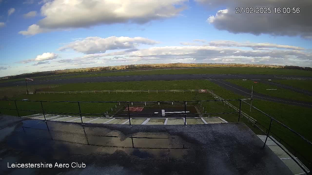 A view from an elevated position overlooking an airfield. The foreground shows a railing, and beyond it, there is a grassy field with a strip of pavement. The background features a cloudy blue sky with some patches of sunlight breaking through. In the distance, there are tree lines and fields. The scene is peaceful with no visible aircraft or vehicles.