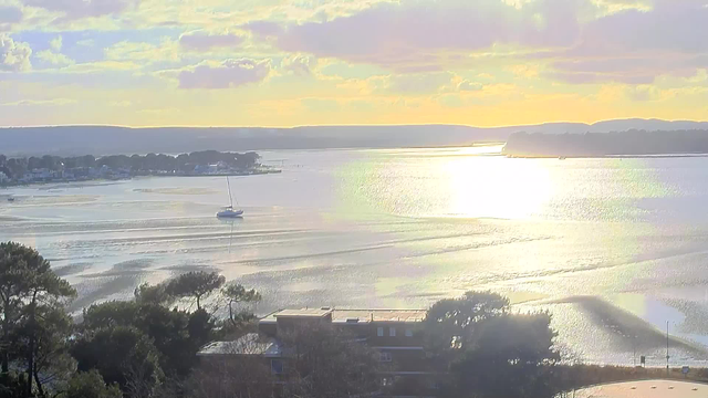 A serene view of a waterway at sunset. The water is shimmering with golden and silver reflections, while a small sailboat drifts near the center. On the left, there are buildings along the shore, surrounded by trees. In the background, gentle hills rise, and the sky is filled with soft pastel clouds, hinting at the fading light of day. The atmosphere is calm and tranquil.