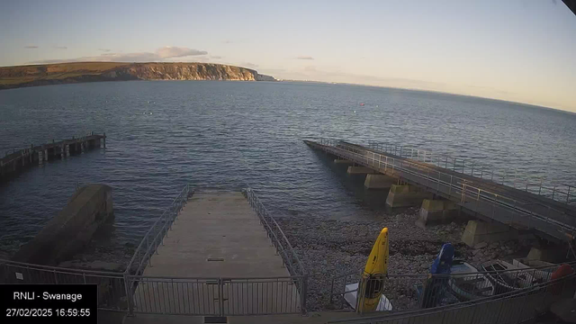 A view of a coastal area with calm blue waters and a shoreline. In the foreground, there is a concrete ramp leading to the water. To the left, a wooden pier extends into the water. Colorful kayaks are stored on the pebbled beach, including a yellow kayak. In the background, cliffs rise up against a clear sky. The scene is peaceful, suggesting a late afternoon atmosphere.