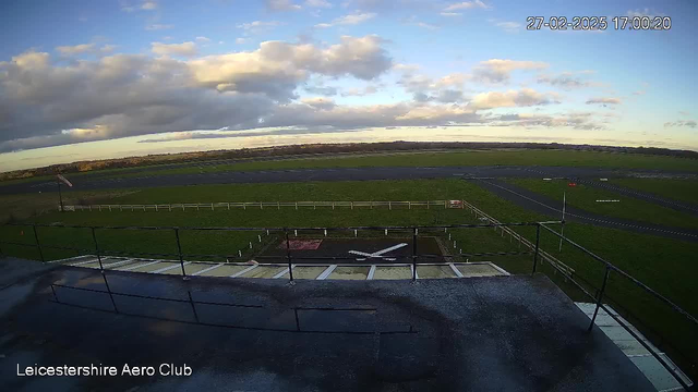 A view from a webcam at Leicestershire Aero Club shows a grassy airfield bordered by a white fence. In the foreground, there is a small aircraft taxiway marked with a white cross, and a few scattered objects. The background features a large blue sky with patchy clouds and a glimpse of distant trees and fields. The lighting suggests it is late afternoon. A flagpole is visible to the left, with a flag gently waving in the wind.