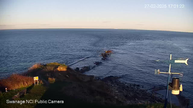 A coastal scene is captured with a view of the ocean extending to the horizon under a clear sky. The foreground includes rocky terrain and patches of green grass, with a wooden bench visible on the left. There is a weather vane and a weather station on the right. A rocky outcrop juts into the water, and small waves create ripples on the surface. The scene is peaceful, with no visible boats or people.