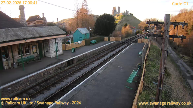 A railway station scene at sunset. The platform is made of stone and features a green bench on the left, a sign indicating "WAY OUT," and a small building with large windows. In the background, there is a greenish-blue shed and a wooden fence. The railway tracks run parallel to the platform, leading towards a hill with ruins on top, partially illuminated by the evening light. Trees and bushes surround the area, creating a natural backdrop.