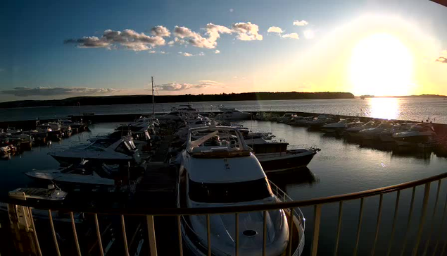 A scenic view of a marina at sunset, showcasing numerous boats and yachts docked in the water. The golden sun sits low on the horizon, casting a warm glow across the scene. There are fluffy clouds scattered in the sky, and the calm water reflects the light from the sun. In the background, a wooded area is visible along the shoreline. The foreground features a railing, indicating the viewpoint is from a higher location.