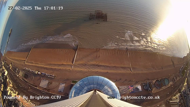 Aerial view from a tall structure overlooking a sandy beach and the sea. The beach is bordered by a pier extending into the water. Waves are gently crashing onto the shore, and there are scattered debris and structures in the water. The foreground shows the rounded rooftop of the building, which is reflective, and the time and date are displayed in the top left corner. In the distance, the coastline and urban area can be seen with some buildings and recreational areas. The image captures a sunny day with bright reflections off the water.