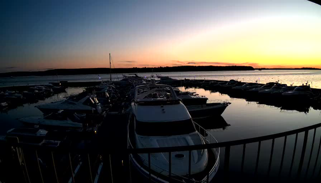 A serene marina at sunset, featuring numerous boats moored in the still water. The sky displays a gradient of colors, transitioning from deep blue at the top to warm oranges and yellows near the horizon. The water reflects the colors of the sky and silhouettes of boats, creating a peaceful atmosphere. A railing is visible in the foreground, adding depth to the scene.