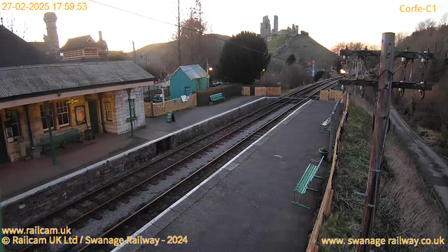 An image of a railway station in Corfe, with a stone building featuring a sloped roof on the left. The foreground includes a platform with several green benches lining it. In the background, there is a hill crowned by a historic castle ruin. The sky is softly illuminated, indicating sunset. Visible are railway tracks leading into the distance. A wooden fence separates part of the platform from a pathway on the right, where power lines and a pole are positioned.