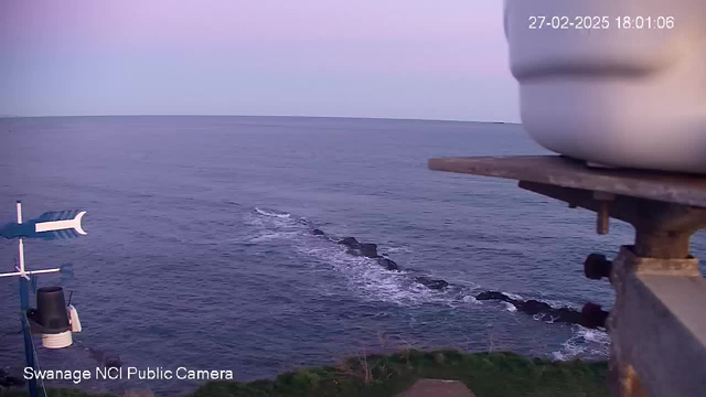 A coastline view showing the ocean and a rocky outcrop extending into the water. The sky has a soft pink hue, indicating sunrise or sunset. In the foreground, part of a weather station is visible, including a wind vane and a bell-shaped structure. The scene captures calm waters with gentle waves, creating a serene atmosphere.