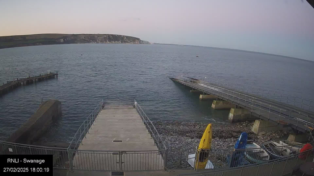 A coastal scene featuring calm blue water extending to the horizon. There are several boats moored at a wooden pier that extends into the water. To the left, a concrete ramp leads down to the water's edge. In the foreground, there are yellow and blue kayaks parked on the shore, surrounded by smooth pebbles. The background features a gentle slope with green hills and cliffs under a pastel sky, indicating early evening light.