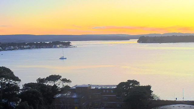 A serene landscape featuring a calm body of water reflecting soft pastel colors of a sunset. In the foreground, there are scattered trees and a low building with several windows. A small sailboat is visible on the water, with distant shorelines and gentle hills in the background. The sky transitions from orange to light blue as the sun sets, creating a peaceful ambiance.