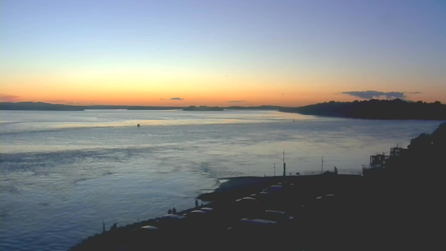 A serene sunset over a body of water, with soft gradient colors ranging from orange to blue in the sky. The water reflects these colors, creating a calm atmosphere. In the foreground, there are silhouettes of cars parked along a shoreline, and a few people are visible, enjoying the view. The surrounding landscape features distant hills and a slight cloud cover in the sky.