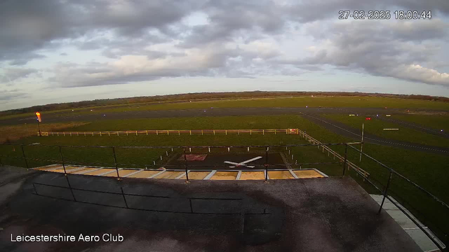 A view from a webcam overlooking Leicestershire Aero Club. The image captures a grassy airfield under a cloudy sky with a few patches of blue. In the foreground, there is a railing and part of a building, while a large X-mark is visible on the ground, indicating a landing area. Various colored lights are scattered around the airfield, and the horizon shows distant trees. The time stamp in the corner indicates the date and time.