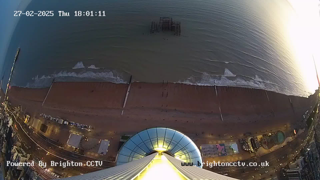 Aerial view looking downward from a tall structure overlooking a sandy beach and the sea. The beach features a wooden pier extending into the water, with some waves breaking onto the shore. The lower portion shows the circular base of the structure, illuminated in yellow, with various buildings lining the edge of the beach. The sky is transitioning from daylight to dusk, with soft colors in the horizon. A date and time stamp is visible in the upper left corner.