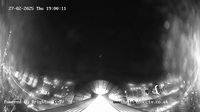 A black and white webcam image shows a nighttime view of a coastal area. The foreground features a curved structure leading towards the center, possibly a roof or a walkway. Below, there are various glowing lights marking a waterfront and some indistinct shapes, likely buildings or attractions. The top of the image is mostly dark, indicating the night sky, with a timestamp showing the date and time. The left side contains a line of road illuminated by lights, leading toward the bottom of the image.