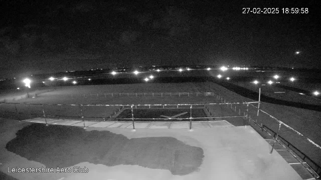 A night view from a webcam at the Leicestershire Aero Club, showing a dark landscape with scattered lights in the distance. In the foreground, there is a flat surface with shadows and a fence partially enclosing an area. The sky is mostly dark with some visible clouds, and the timestamp reads 18:59:58 on February 27, 2025.