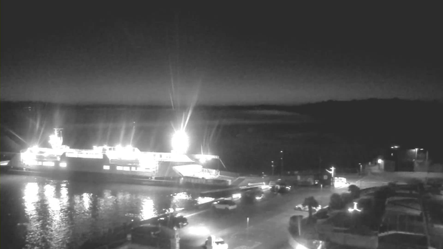 A dimly lit scene at night featuring a large ferry docked at a pier. The ferry is illuminated with bright lights, reflecting on the water's surface. In the foreground, there are some vehicles parked along the waterfront, and faint outlines of trees and buildings can be seen near the shore. The sky is dark, with a hint of light from the horizon.