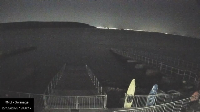 A dark scene showing a waterfront area at night. In the foreground, there are two kayaks, one yellow and one blue, positioned next to a railing. The water appears calm, and in the distance, faint lights can be seen along the horizon, possibly from a town or city. The image is low in light, creating a moody atmosphere.