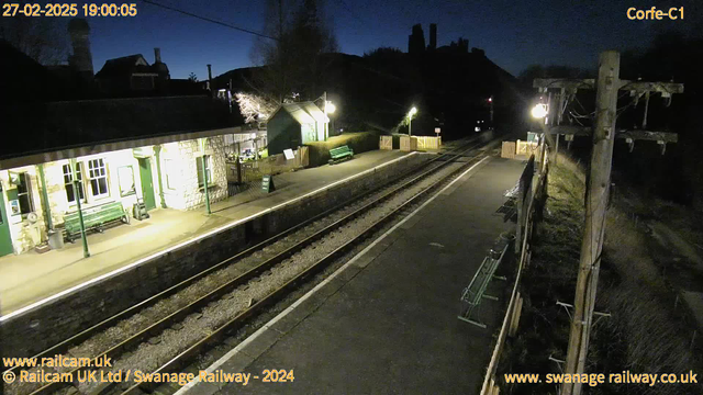 A railway station at dusk. The platform is mostly empty, with two green benches on the left side. A building made of light-colored stone is on the left, featuring several windows and a sign indicating the exit. There are lights illuminating the area, giving off a warm glow. Tracks run alongside the platform, leading into the distance. In the background, there are dark silhouettes of the hills or buildings. A power pole with wires stands to the right. Overall, the scene has a calm, quiet atmosphere.